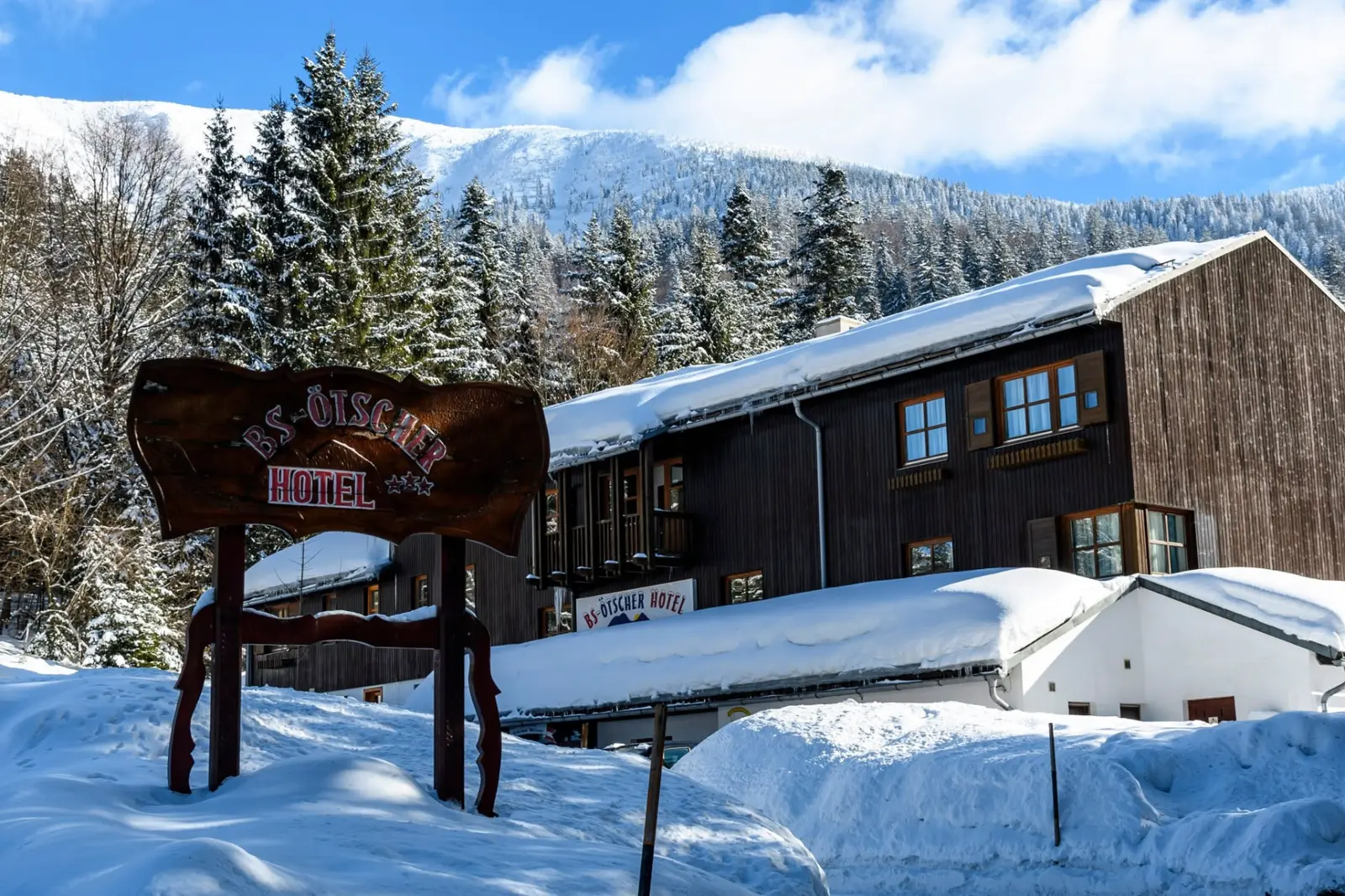 A BS-Ötscher Hotel fa cégtáblája a havas fenyvesekkel és az Ötscherrel a háttérben